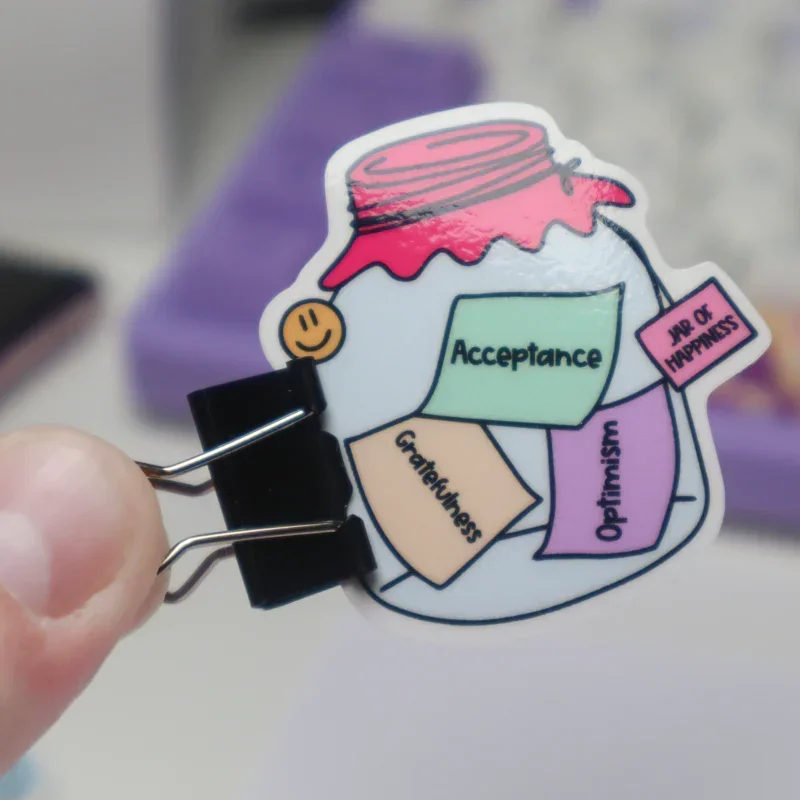 Kuvassa on laminoitu tarra "Jar of Happiness" eli onnellisuuden purkki. Tarra toimii kalentereissa, juomapulloissa, läppäreiden kansissa ja vaikka puhelimien kuorissa hyvänä muistutuksena ja motivaatiolauseena. Purkin sisällä on muistilappuja, joissa lukee hyväksyntä, kiitollisuus ja optimismi. Tarra on valmistettu Suomessa laadukkaista materiaaleista. Laminointikalvo on kotimaiselta valmistajalta ja PP-tarramateriaali tulee Euroopan sisältä.