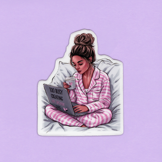 Tarra, jossa tummahiuksinen nainen juo kahvia ja suunnittelee unelmia tietokoneella sängyssä. Naisen hiukset on nutturalla. 